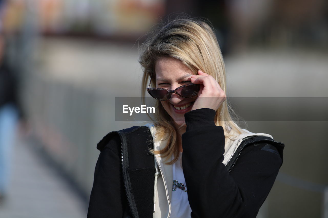 one person, adult, glasses, blond hair, fashion, women, sunglasses, spring, jacket, portrait, hairstyle, focus on foreground, young adult, long hair, clothing, person, front view, business, city, emotion, day, smiling, architecture, happiness, outdoors, human face, leather jacket, lifestyles, standing, female