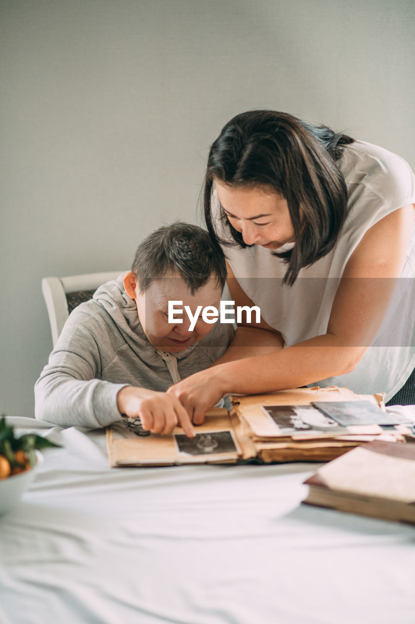 Asian girl points a smiling elderly woman with down syndrome to a photo in an old album.