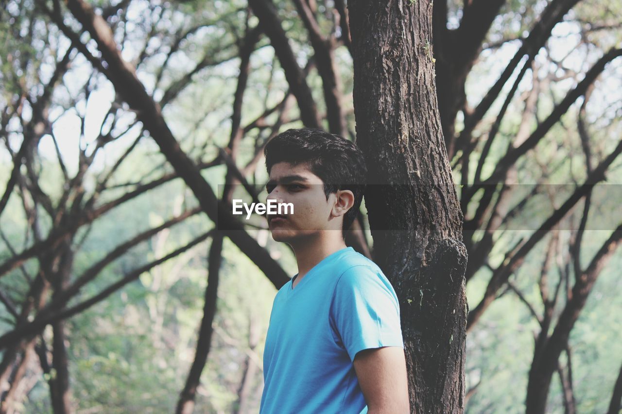 Side view of thoughtful young man standing by tree trunk in forest