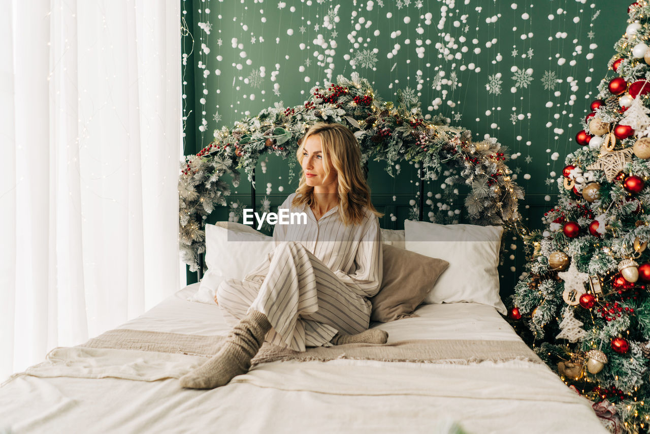 Young beautiful blonde woman in pajamas sits on the bed on a festive christmas morning.