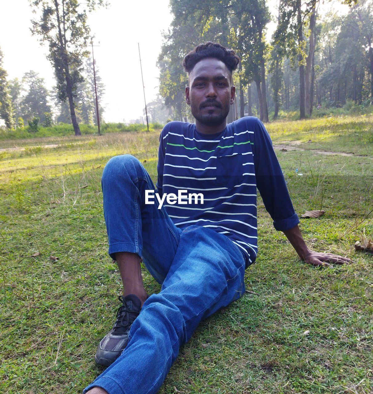 one person, plant, casual clothing, grass, leisure activity, men, nature, looking at camera, front view, lifestyles, portrait, young adult, tree, land, day, adult, full length, sitting, field, clothing, person, relaxation, beard, facial hair, outdoors, smiling, green, meadow, footwear, lawn