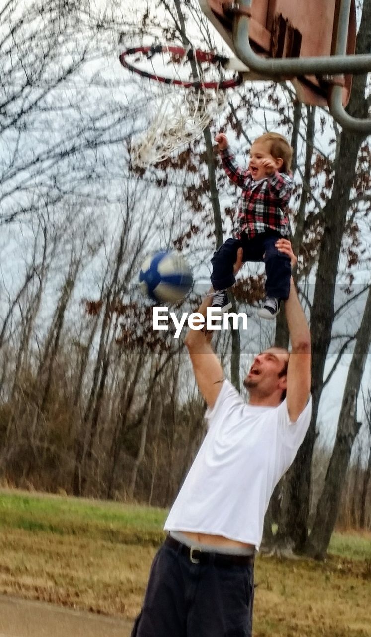 Father carrying baby boy to put basketball in hoop on field