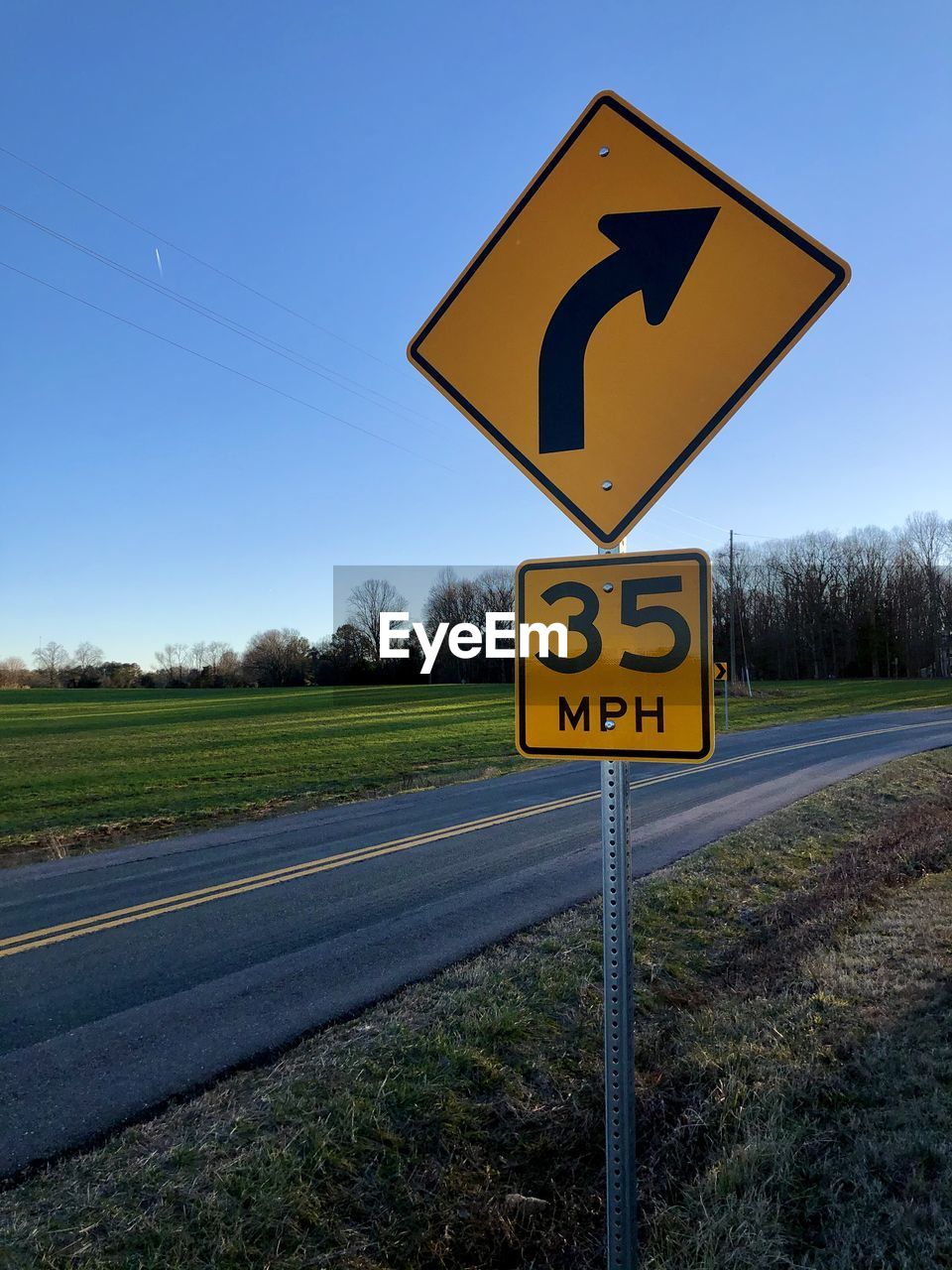 Curve ahead road sign on field against clear blue sky and next to a road with a double yellow line.