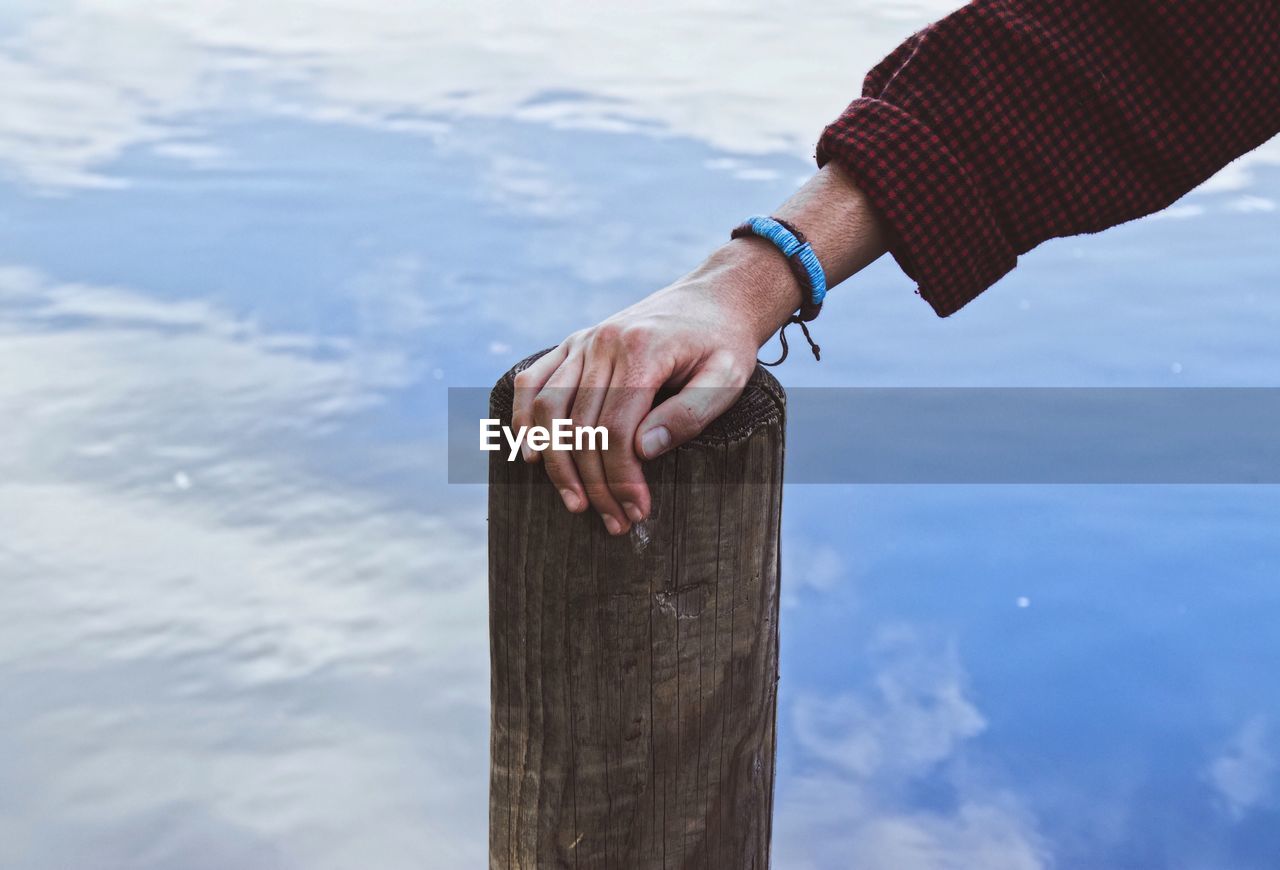 Cropped hand of man touching wooden post in lake