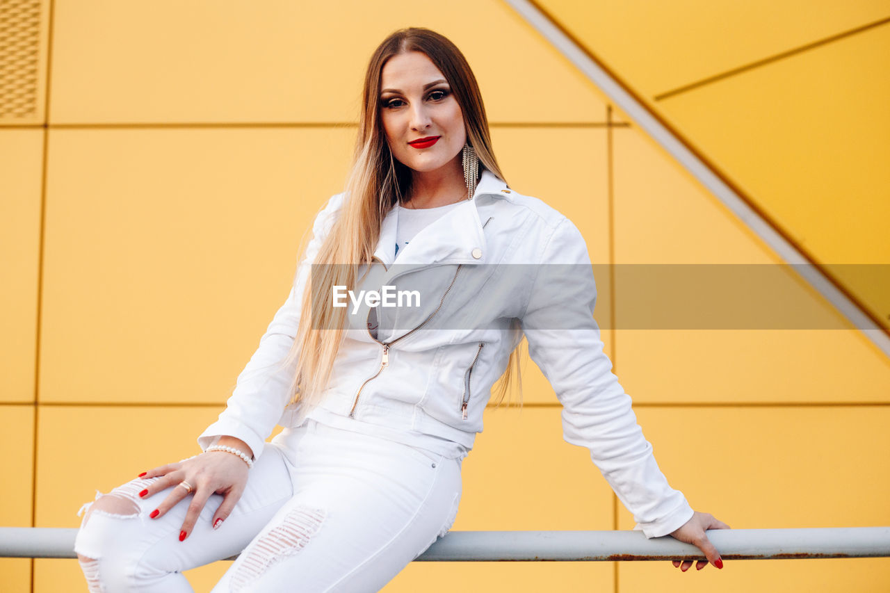 Portrait of young woman sitting on railing against wall