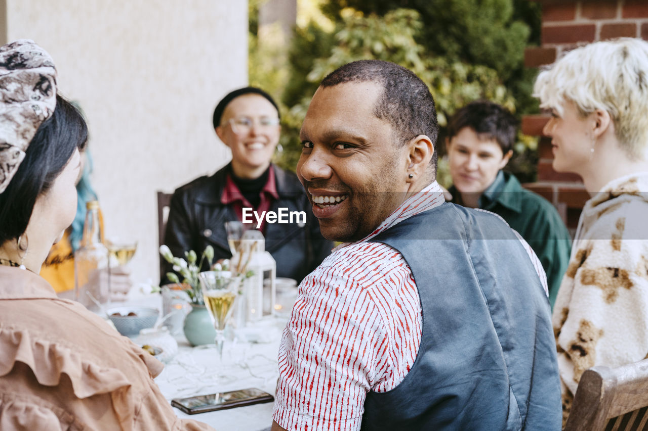 Portrait of smiling gay man looking over shoulder by friends during dinner party in back yard