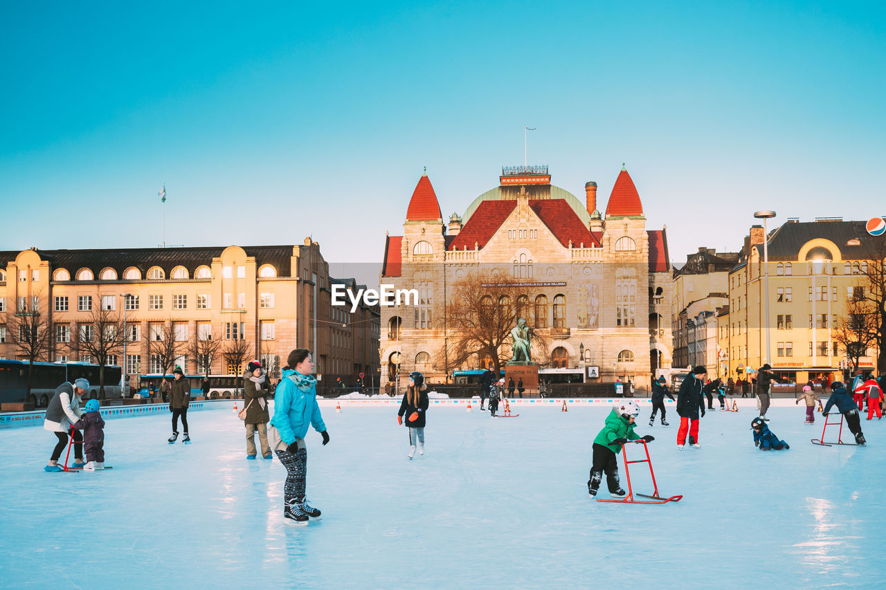 ice skating, architecture, winter sports, ice rink, building exterior, winter, cold temperature, ice, group of people, built structure, travel destinations, city, nature, crowd, snow, sports, sky, building, large group of people, skating, travel, resort, blue, tourism, history, vacation, leisure activity, the past, clear sky, lifestyles, holiday