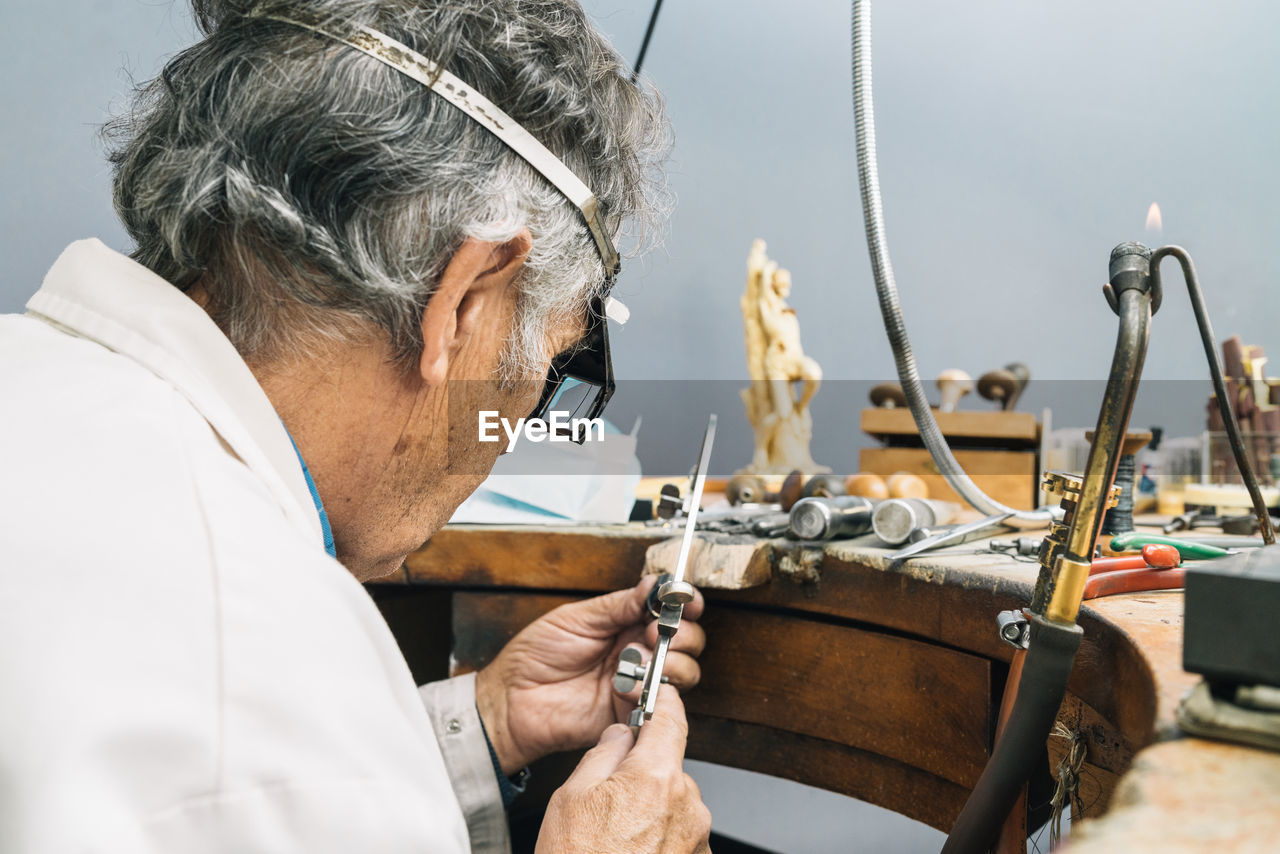 Side view of concentrated skilled mature male master in glasses using ring sizer while creating jewelry in workshop