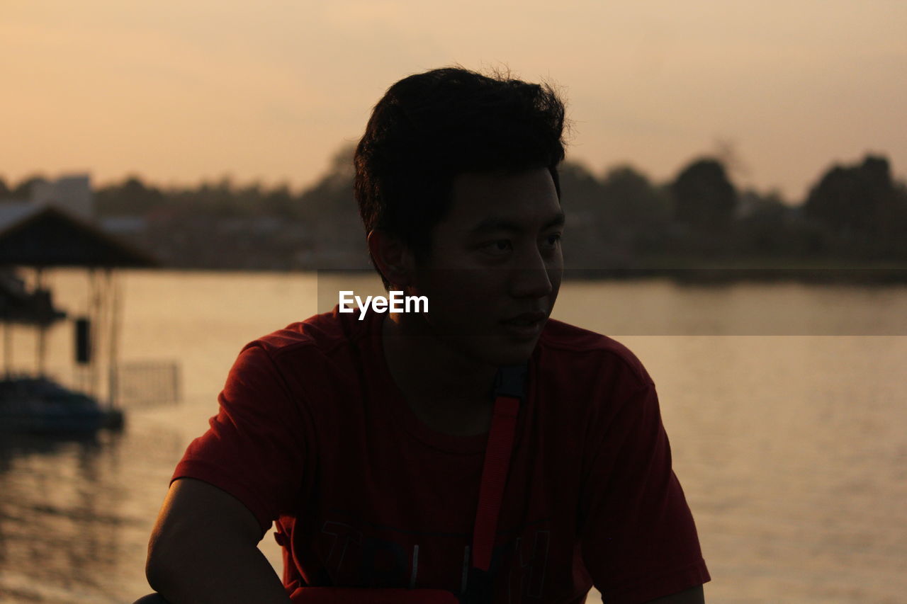 PORTRAIT OF YOUNG MAN STANDING BY LAKE AGAINST SKY