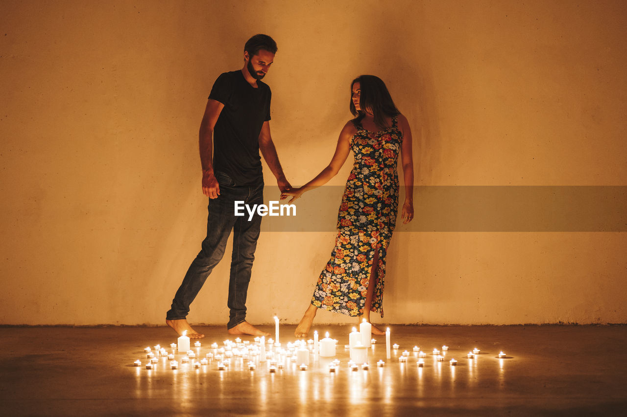 Couple holding hands while standing by lit candles against wall at home