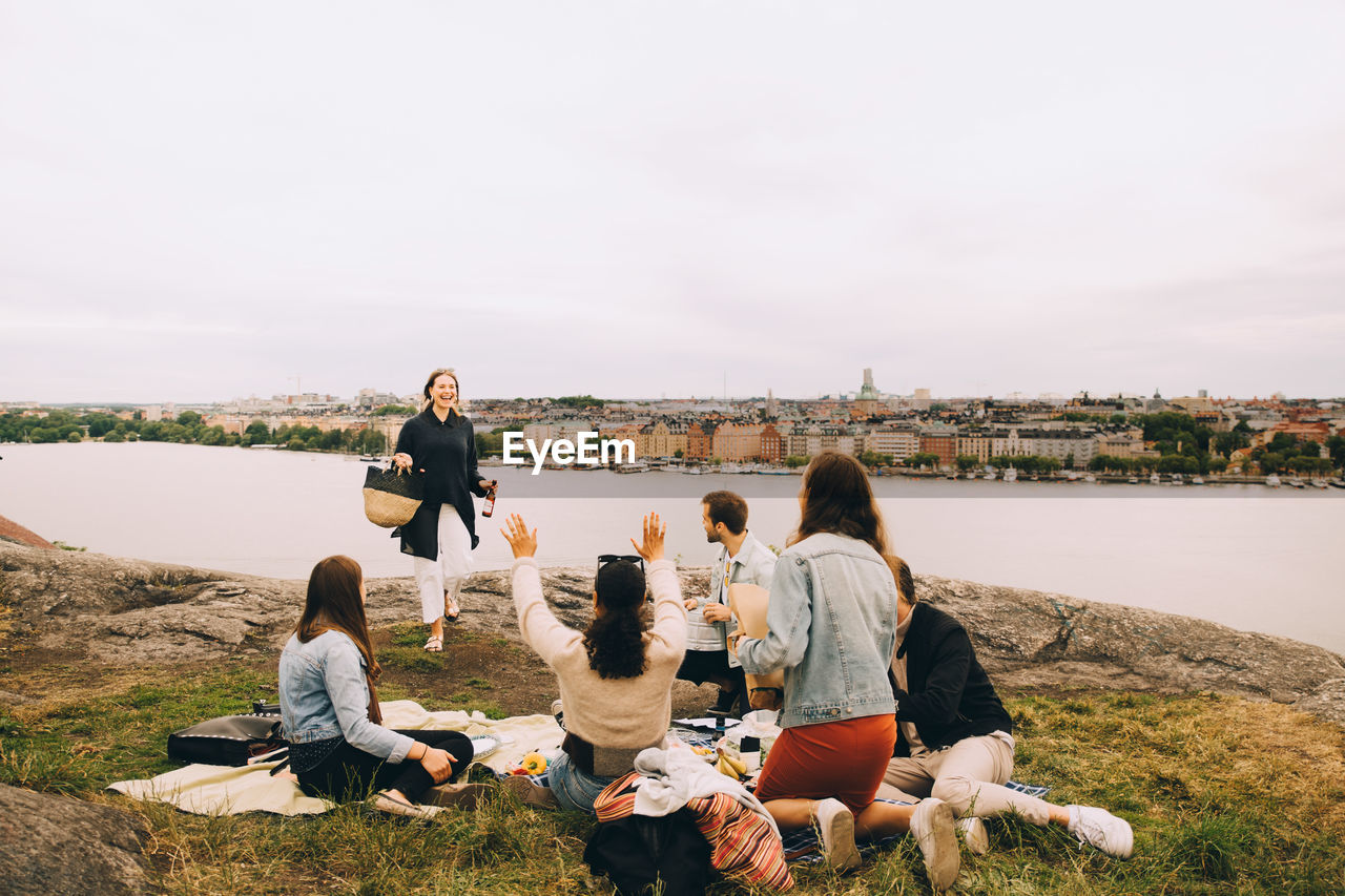 Friends greeting woman walking on field by lake against sky