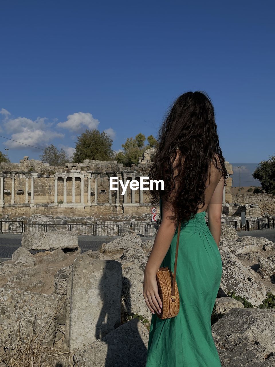 one person, architecture, history, nature, sky, the past, women, vacation, old ruin, ancient, adult, travel destinations, travel, hairstyle, built structure, rear view, long hair, temple, blue, leisure activity, tourism, day, young adult, ancient history, dress, standing, ruins, land, archaeology, brown hair, rock, waist up, person, sunlight, sunny, outdoors, clothing, lifestyles, clear sky, fashion, tourist, ancient civilization, building exterior, three quarter length, trip, casual clothing