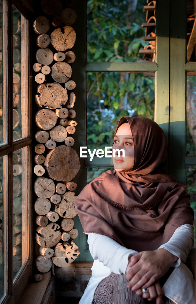 Thoughtful woman wearing hijab looking through window while sitting at home