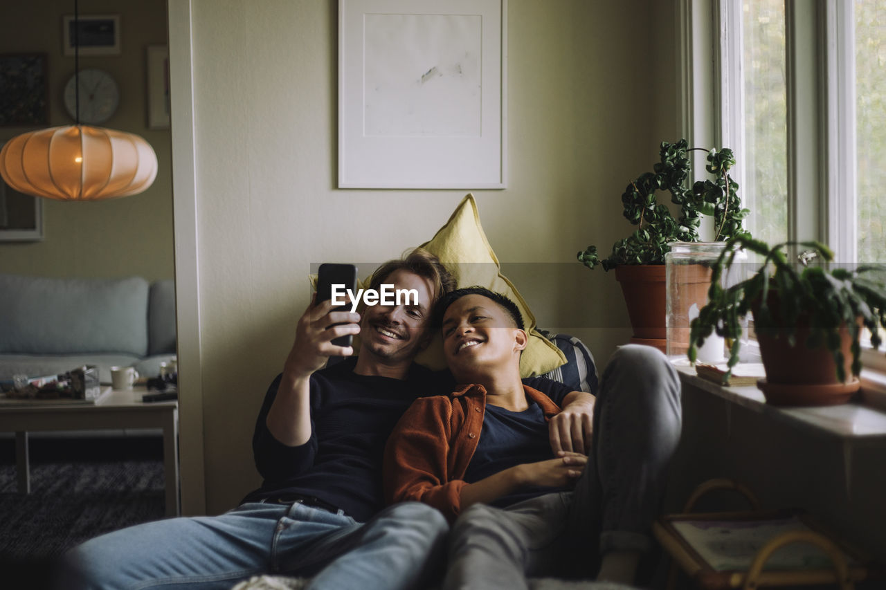 Smiling gay couple sharing smart phone while sitting near window at home