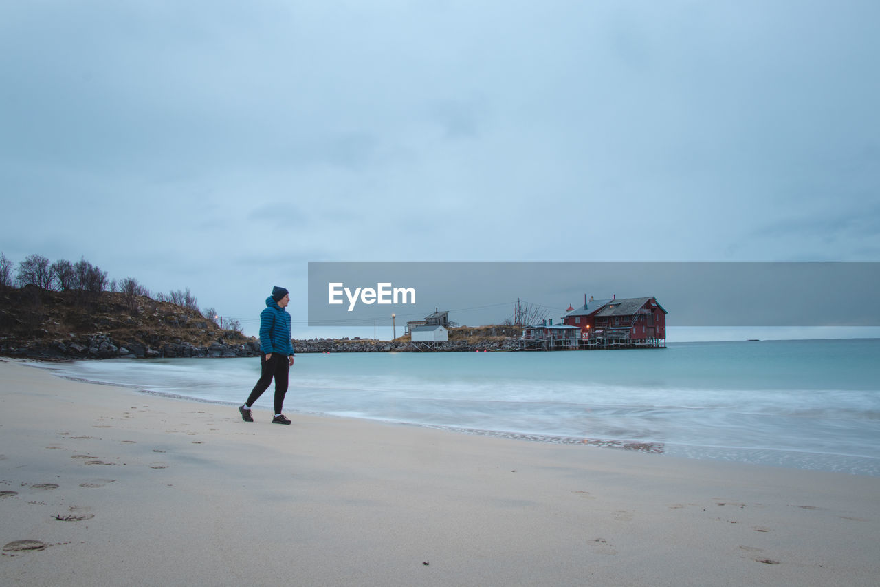 Man in a blue jacket walks along  sandy beach in village of bovaer at the end of senja in norway.