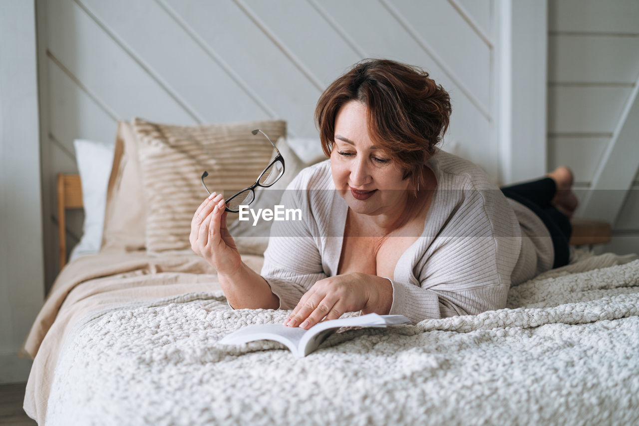 Smiling middle aged plus size woman lying on bed and reading book at home