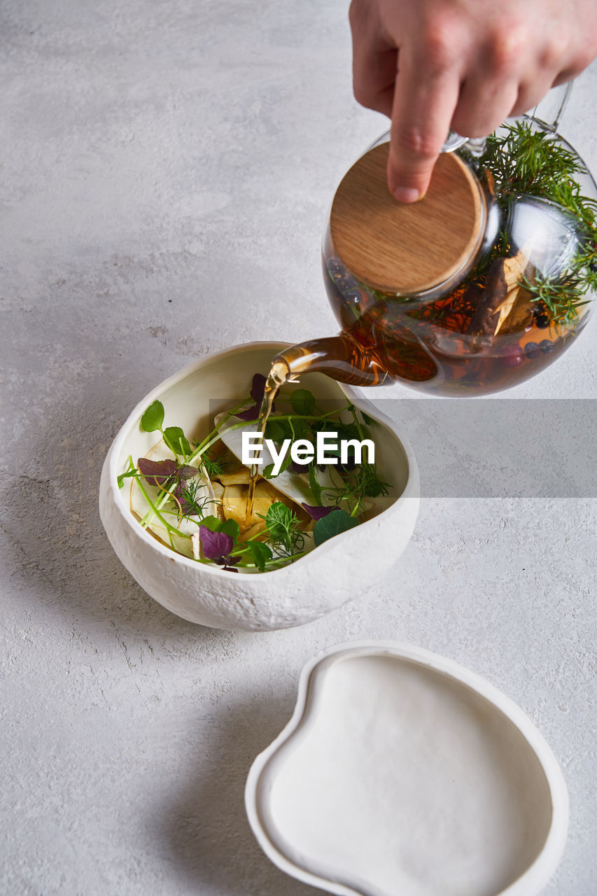 Unrecognizable person pouring fresh warm tea from glass teapot into ceramic bowl with herbs