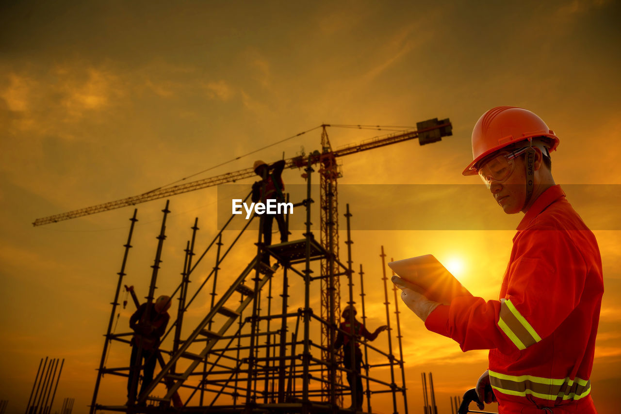 Civil engineer with digital tablet standing near people working on construction site against sky during sunset