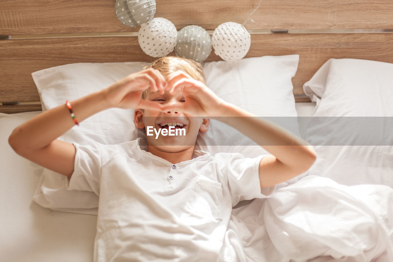 High angle view of cheerful boy making heart shape while lying on bed at home