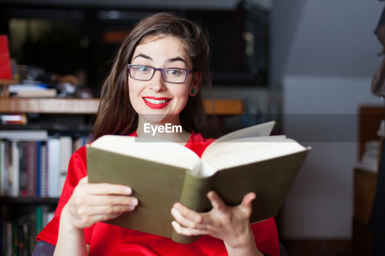 Smiling young woman wearing eyeglasses while reading book at home