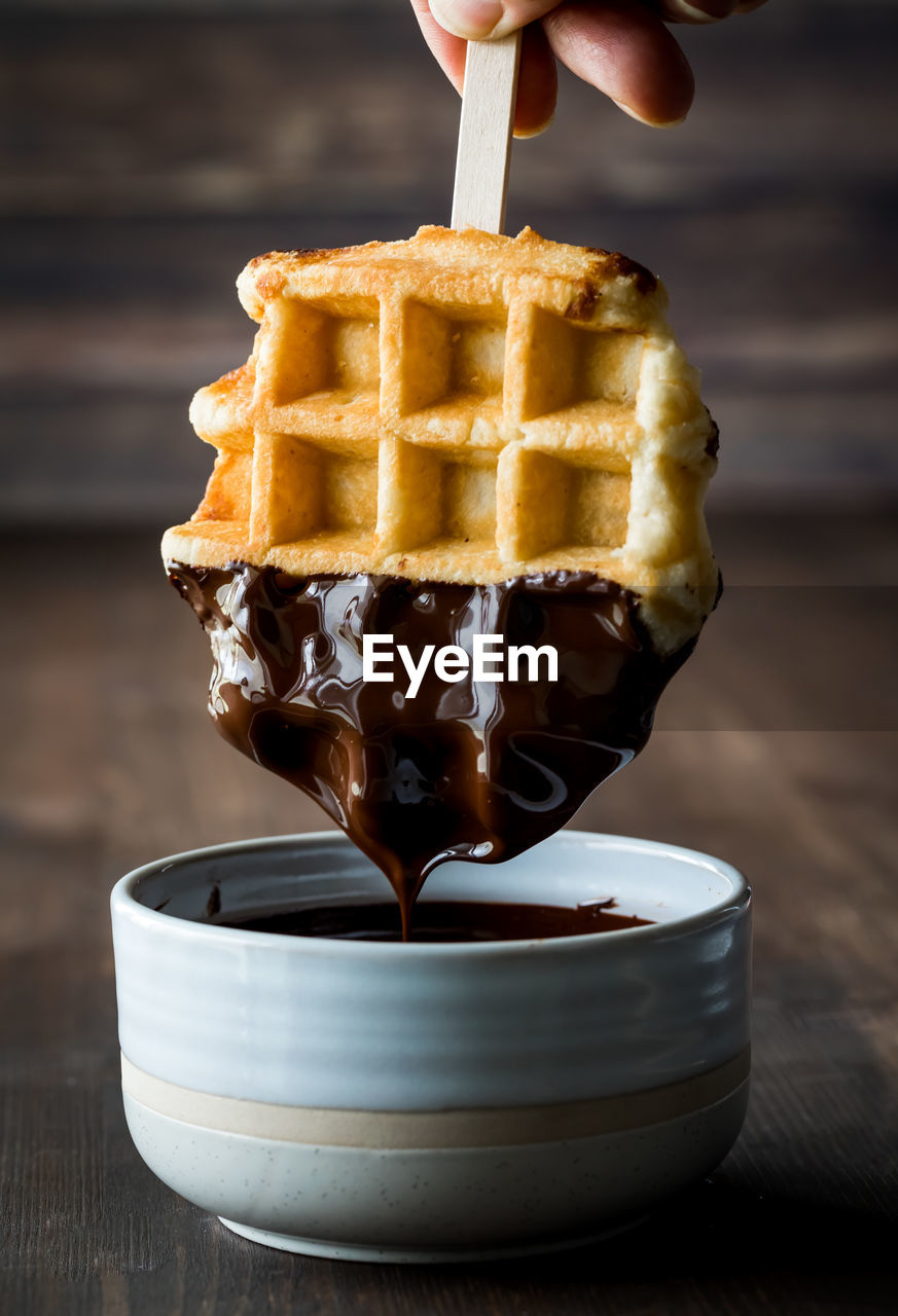 A waffle being dipped into melted chocolate and dripping into a bowl.