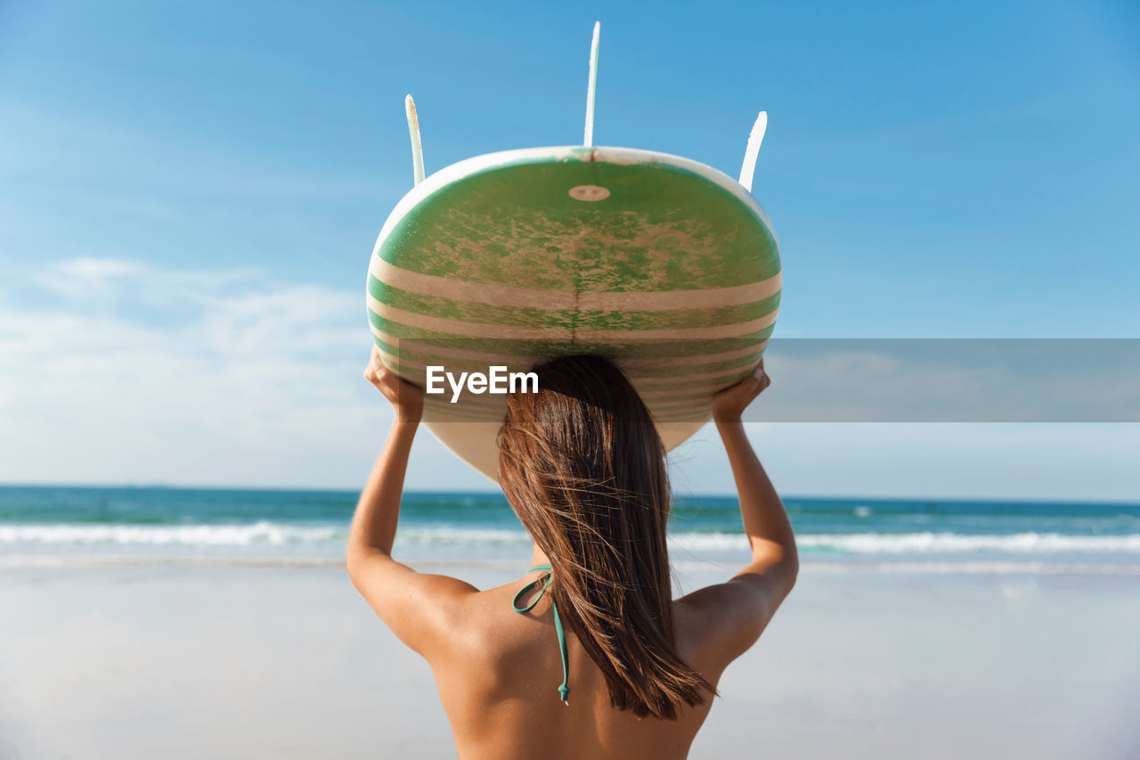 Rear view of young woman holding surfboard while standing at beach against sky