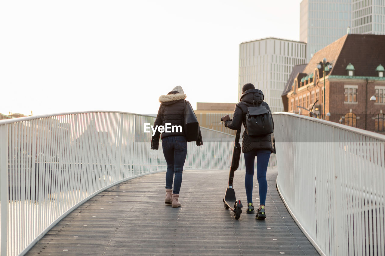 Rear view of female friends walking with e-scooter on bridge in city