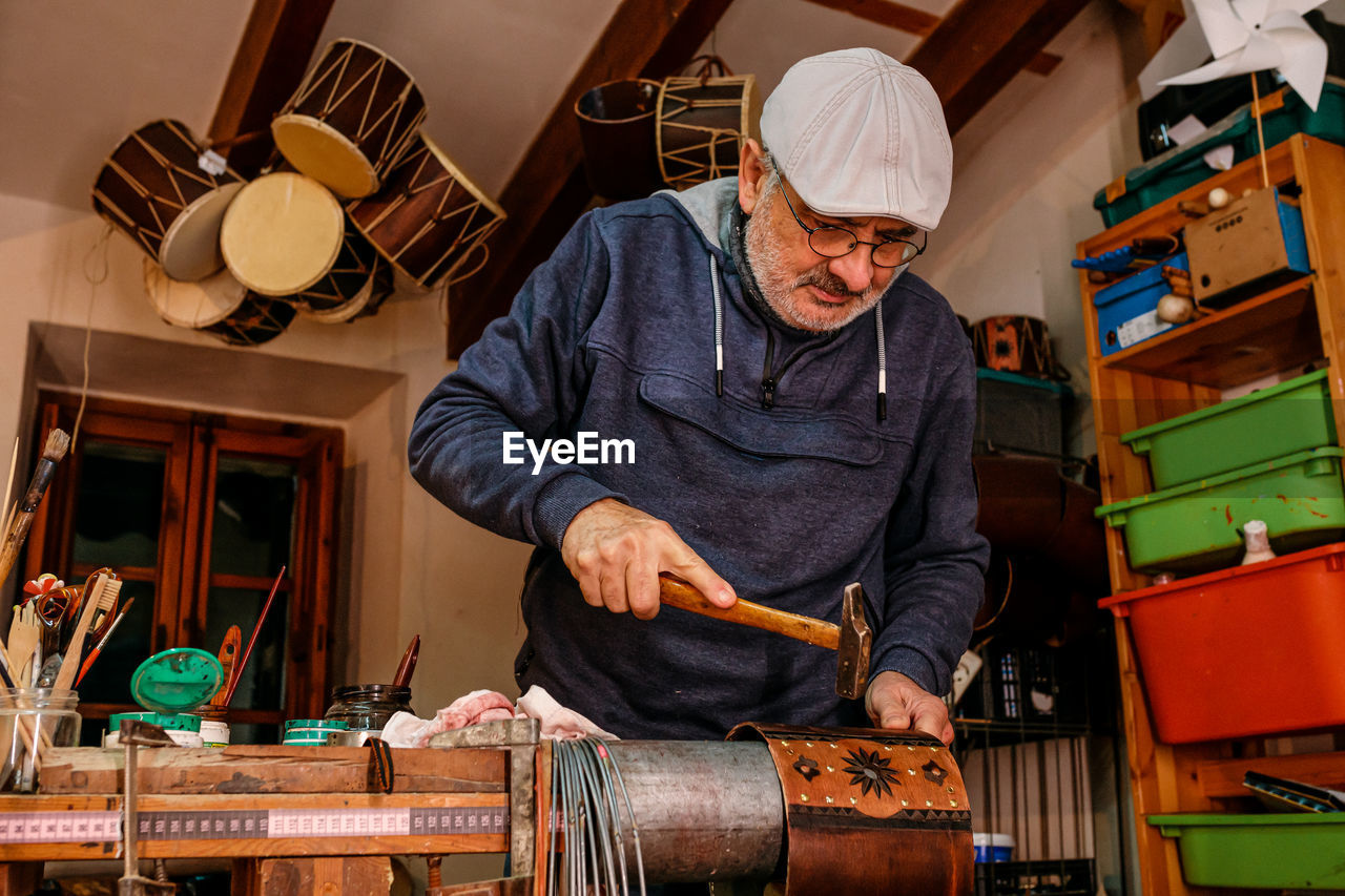 Professional male master using hammer while making shell of drum with creative patterns while working in light workshop with special instruments