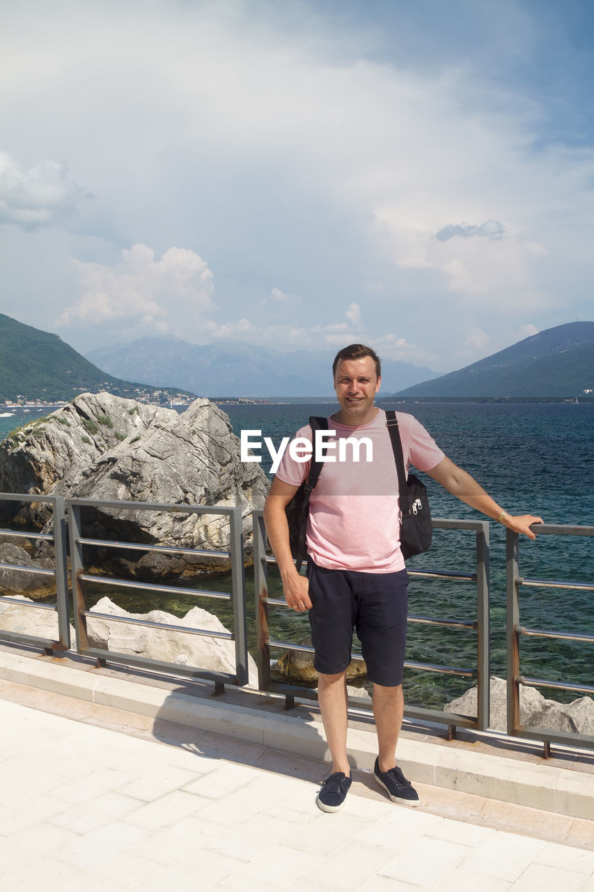 FULL LENGTH PORTRAIT OF SMILING STANDING ON MOUNTAIN AGAINST SKY