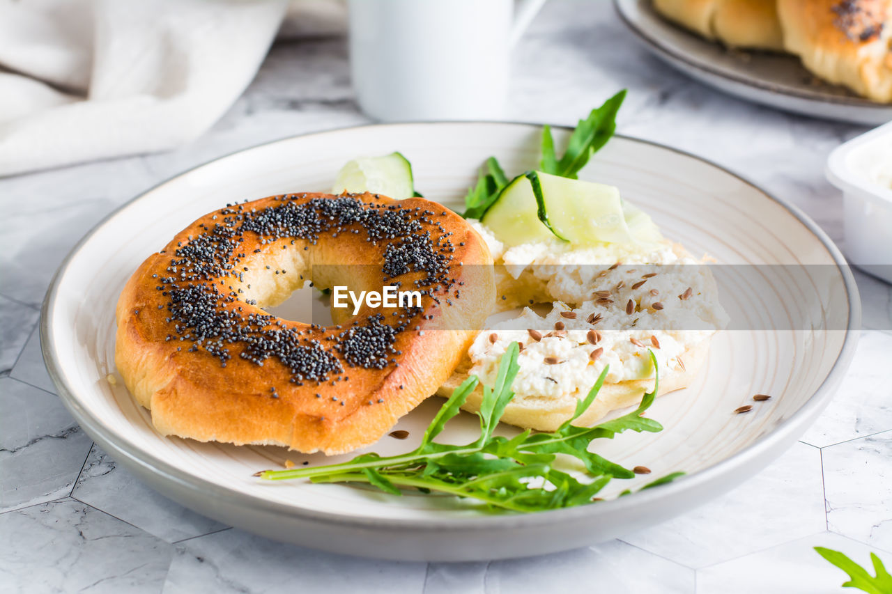Bagel sandwich with poppy seeds with ricotta, cucumber and arugula on a plate on the table. 