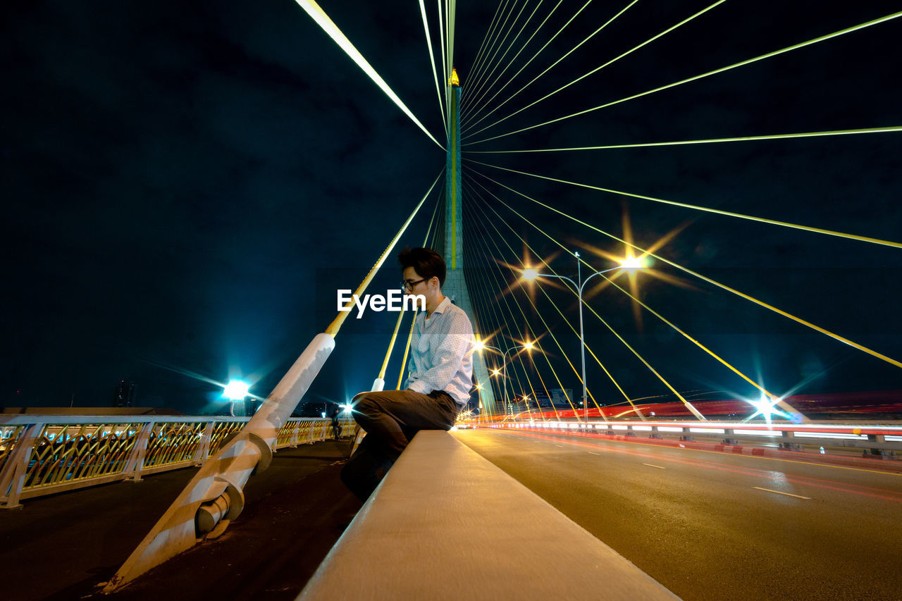 Side view of man sitting on bridge with light trails on road against sky at night