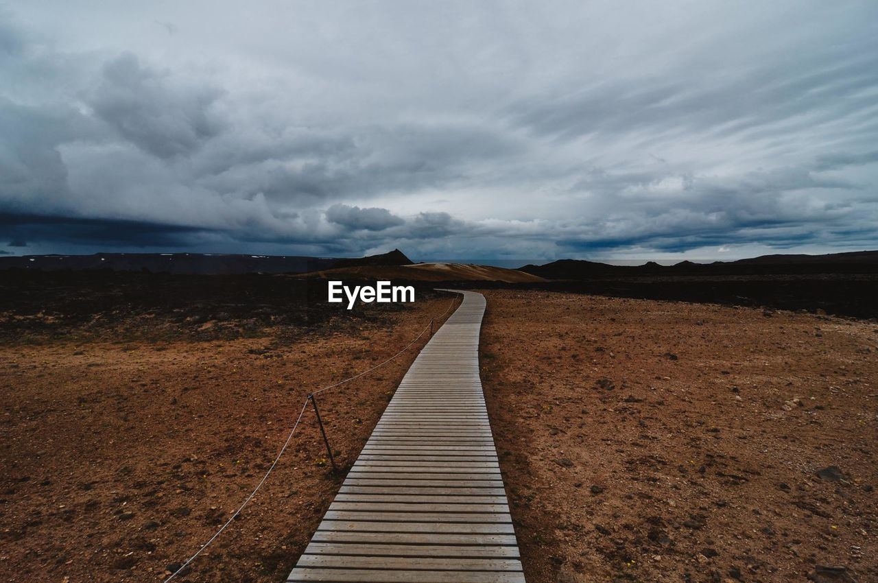 Boardwalk amidst field against cloudy sky