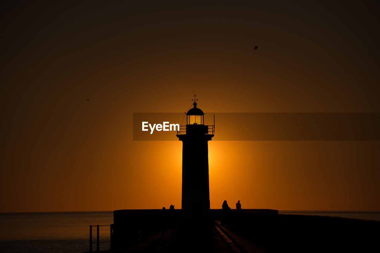 Silhouette lighthouse on beach against sky during sunset