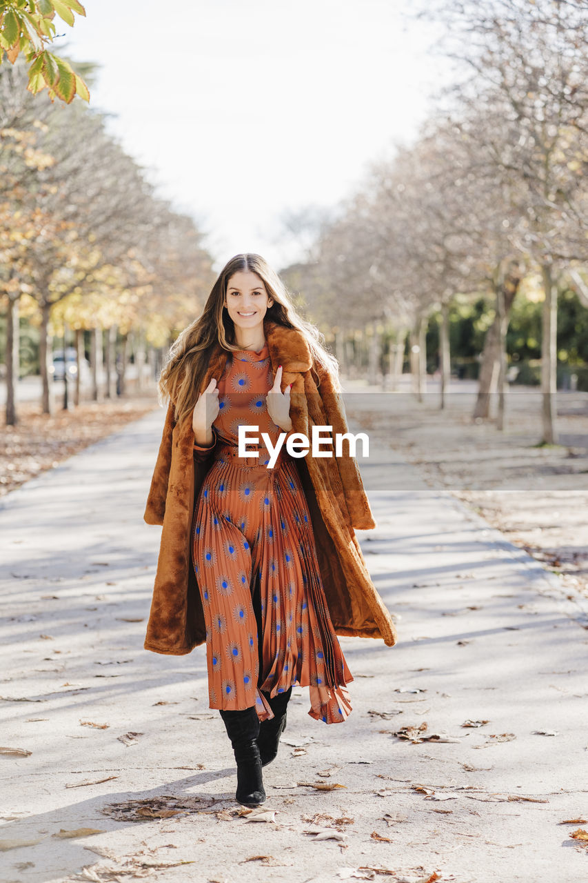 Smiling fashionable woman wearing fur coat walking on footpath during sunny day