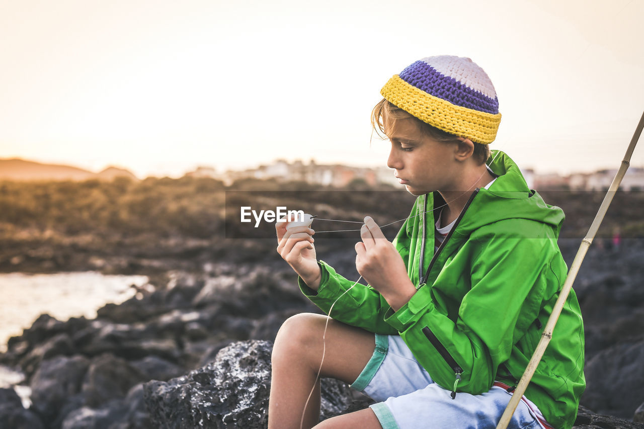 Boy holding fishing rod while sitting against sky