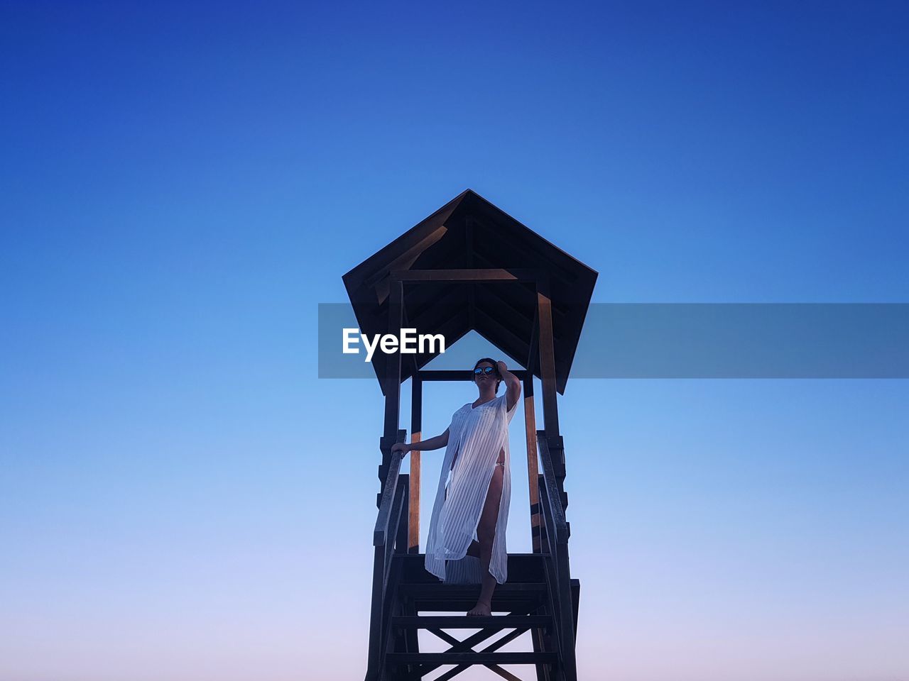 Low angle view of woman standing on lifeguard hut against clear blue sky during sunset