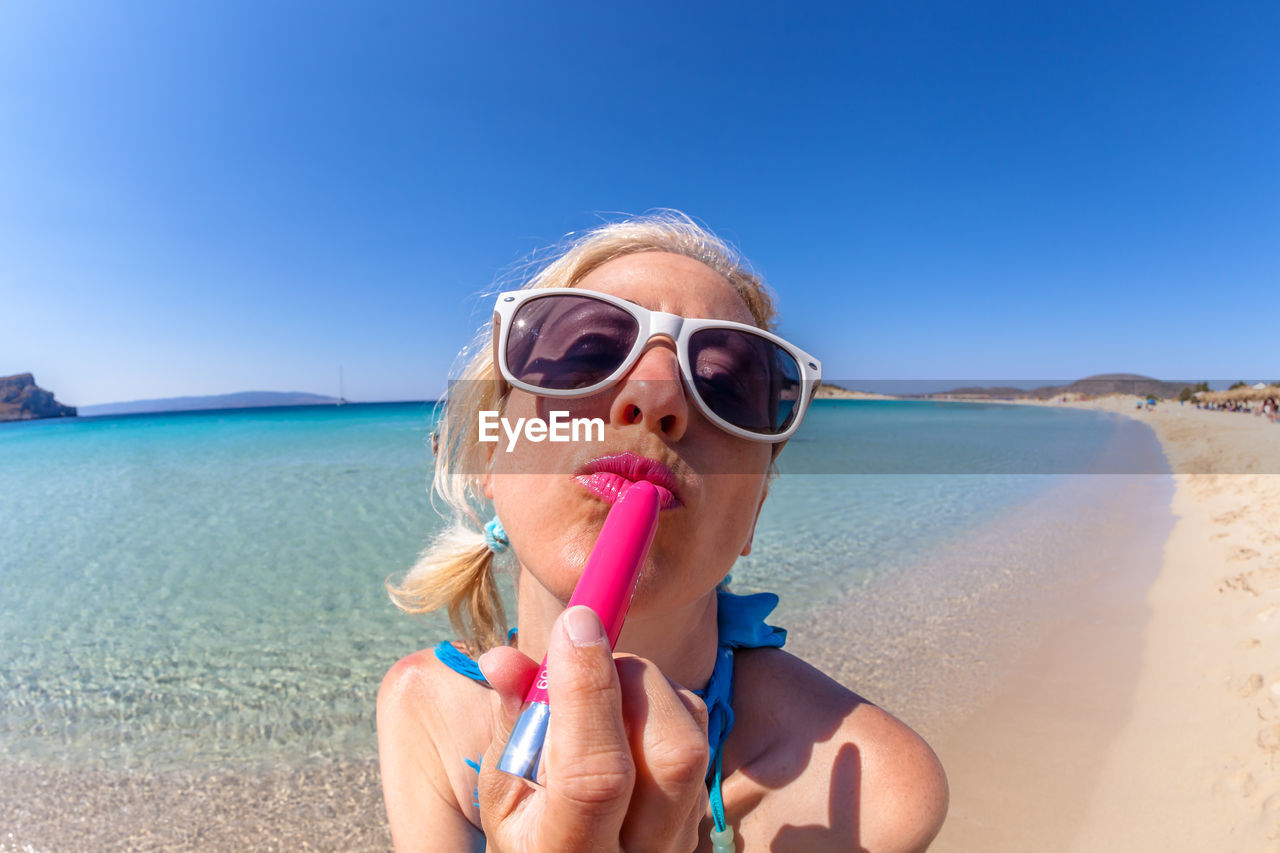 Close-up of mature woman applying lipstick while wearing sunglasses at beach