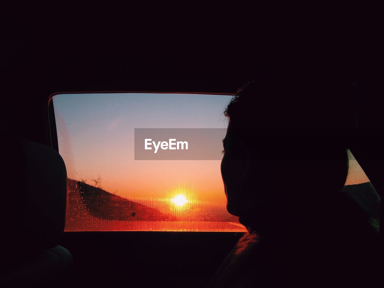 Teenage girl looking through window while sitting in car during sunset