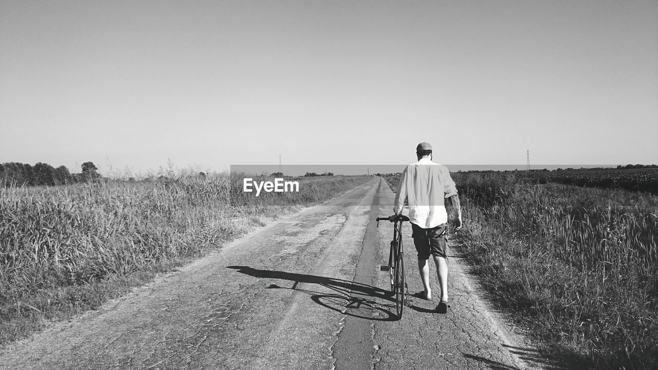 Full length rear view of man with bicycle walking on road against sky during sunny day