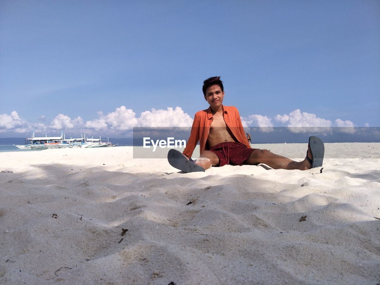 Portrait of young man sitting on sand at beach against sky