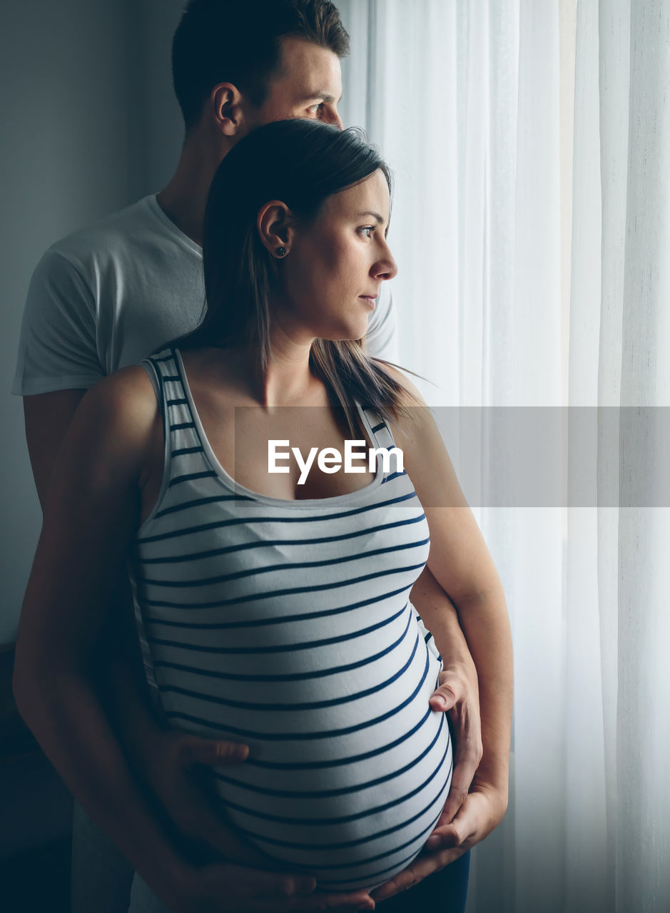 Pregnant woman with husband standing by window at home