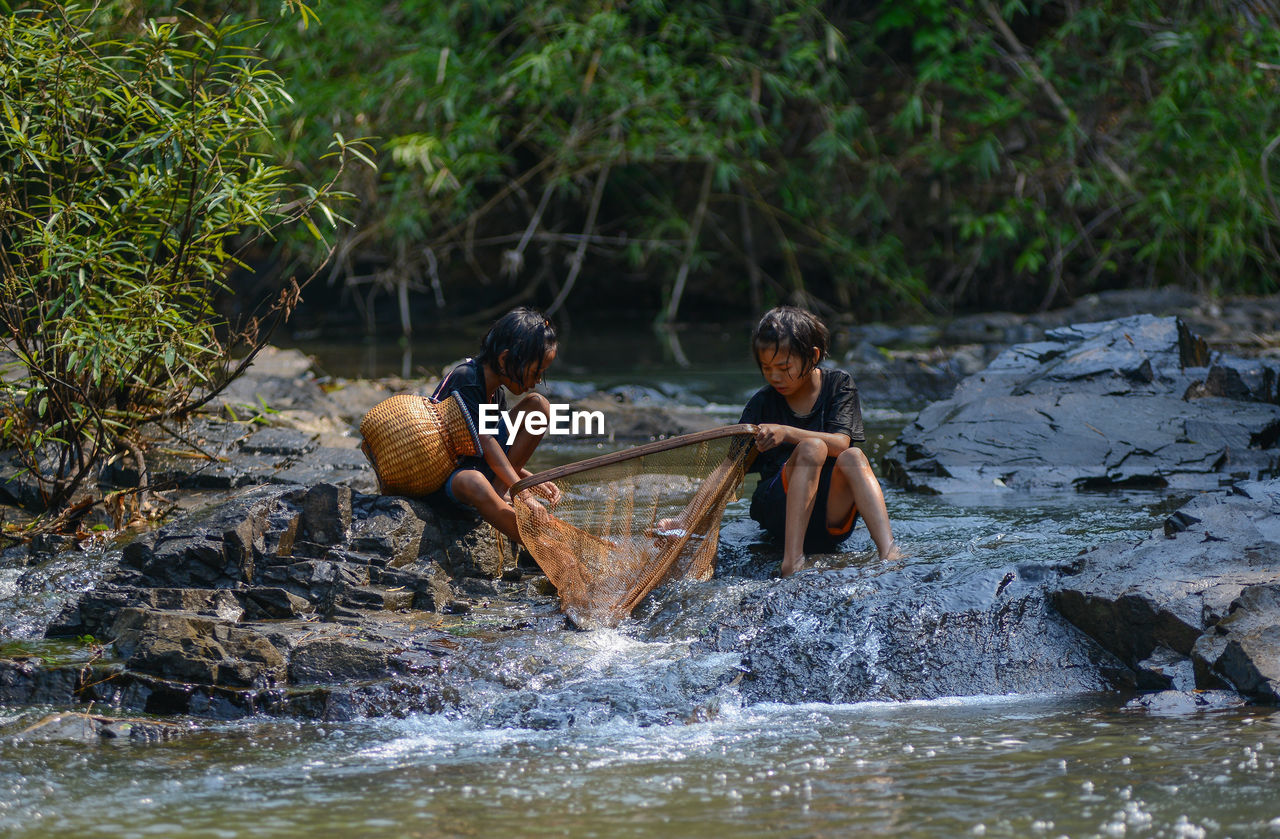 Sisters fishing while sitting at stream in forest