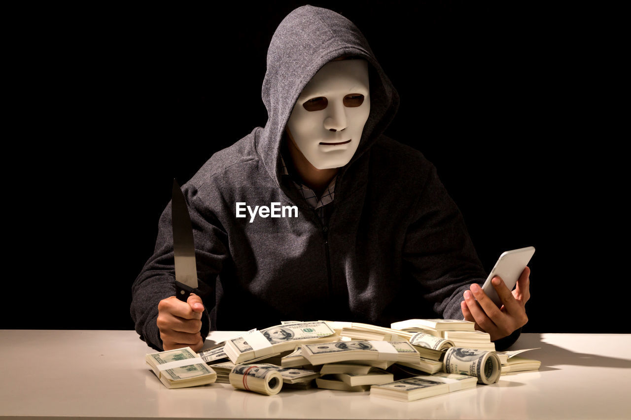 Criminal in mask using mobile phone while sitting with currency at table against black background