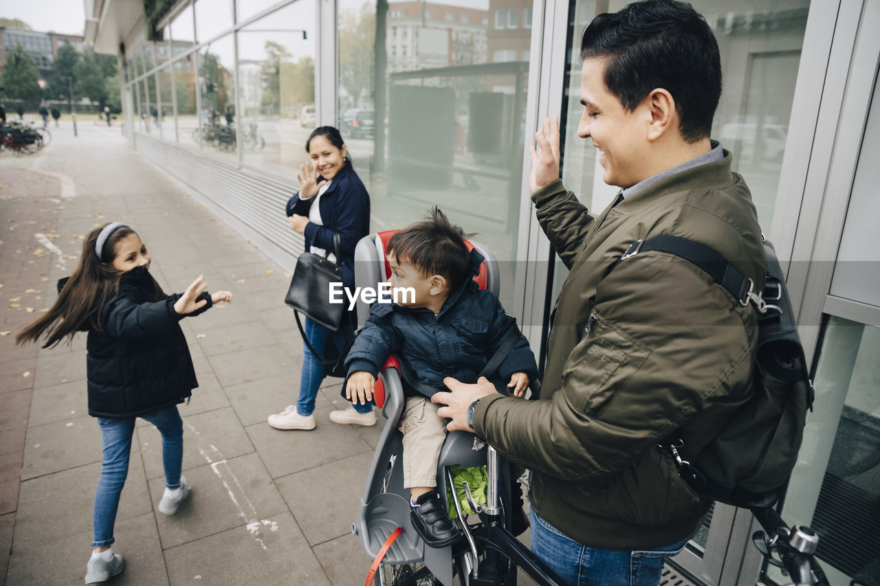 Smiling mother and daughter waving to boy with father on sidewalk in city