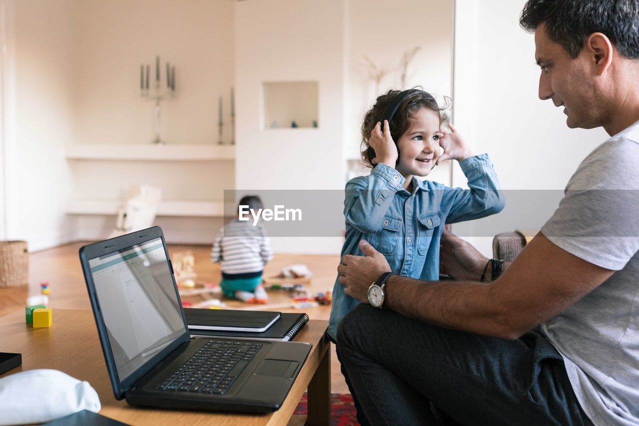 Smiling daughter with headphones standing by father while brother playing in background at home