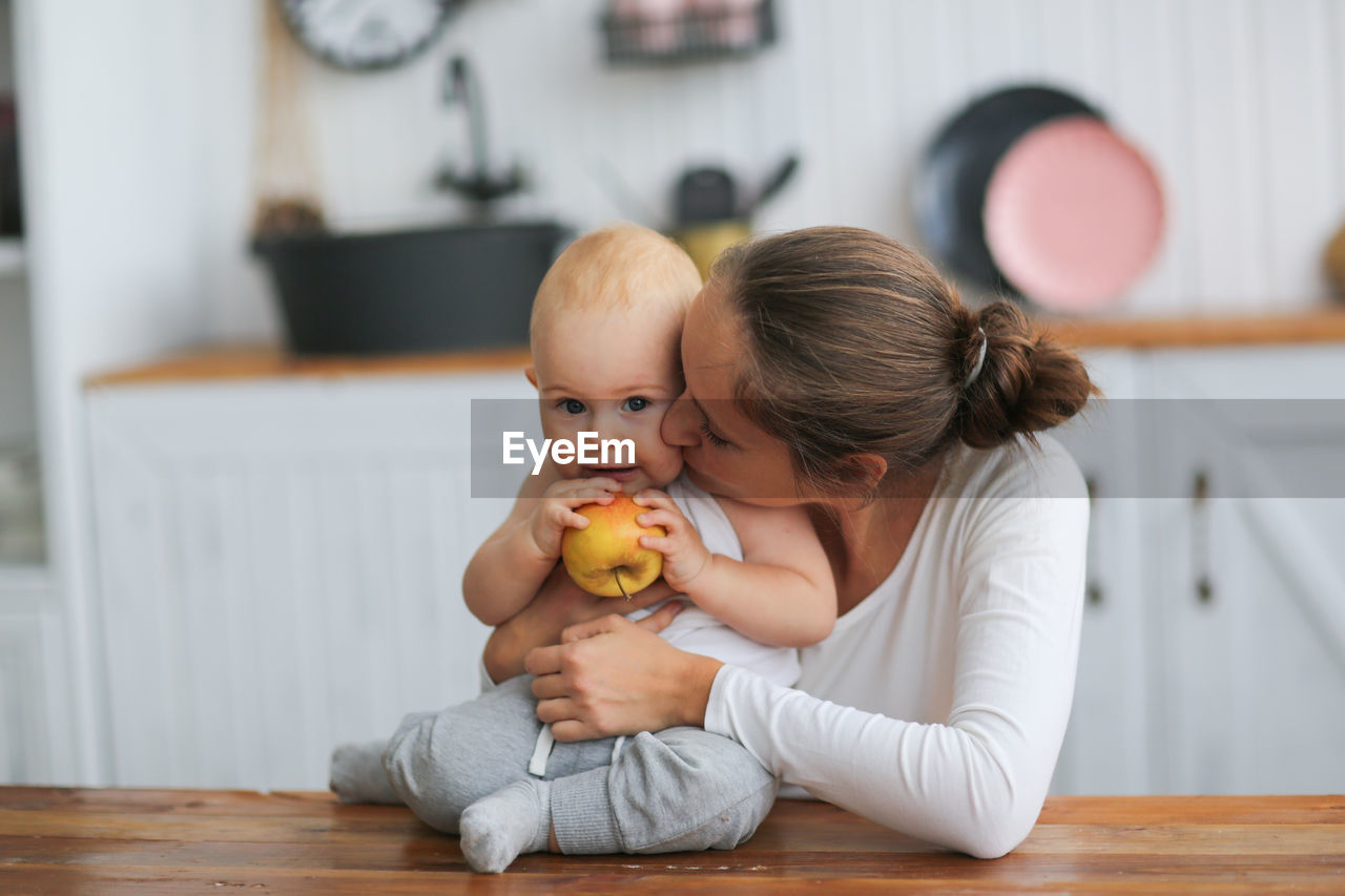 Baby with an apple and caring mother in the kitchen in a real interior.