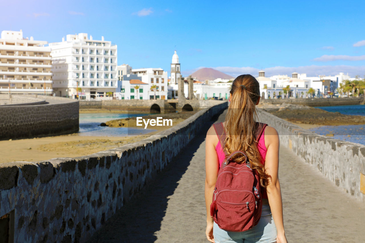 Rear view of woman with backpack walking in city against sky