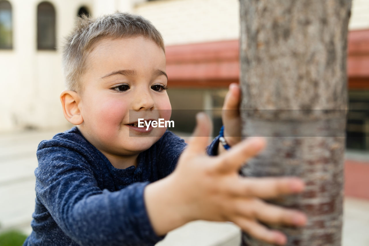 Close-up of smiling cute boy touching tree trunk 