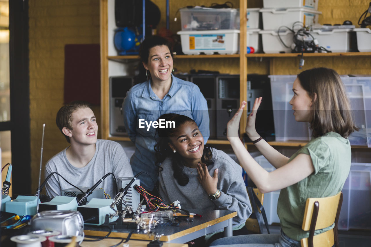 Smiling female student gesturing while sitting with friends and teacher in classroom at high school