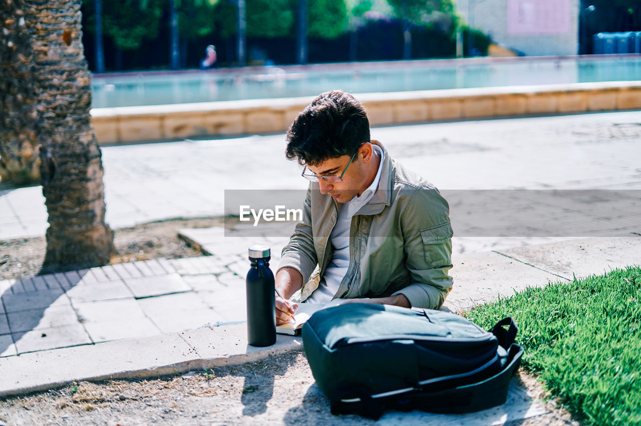 Focused male student sitting on stone bench in university campus and writing in notepad while doing homework on sunny day